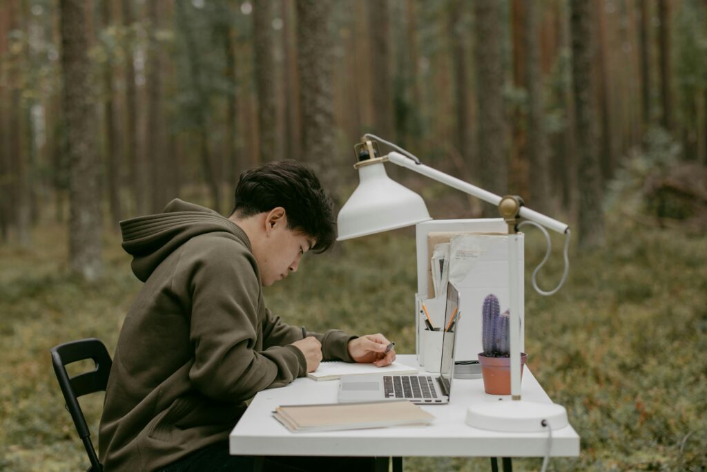 A person studying outside using a laptop and notebook in a natural setting. Ideal for education or remote work concepts.
